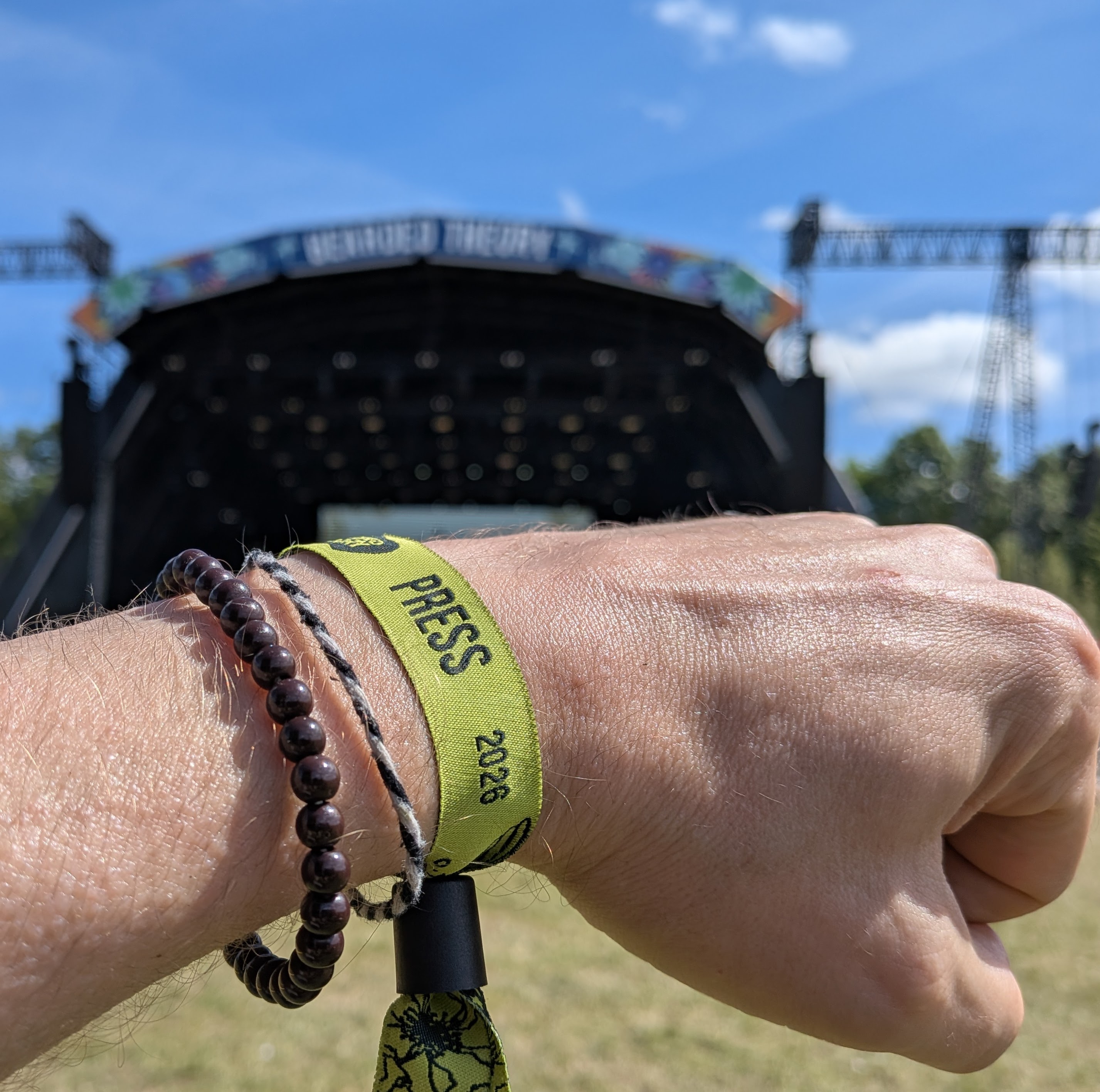 Finbarre Snarey’s green Bearded Theory 2026 press wristband for Tarot Interviews, photographed in front of the festival main stage on a sunny weekend in Derbyshire.