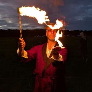 Finbarre Snarey training in fire performance at Wyldwood Renaissance Festival, holding flaming props against a dark evening sky.
