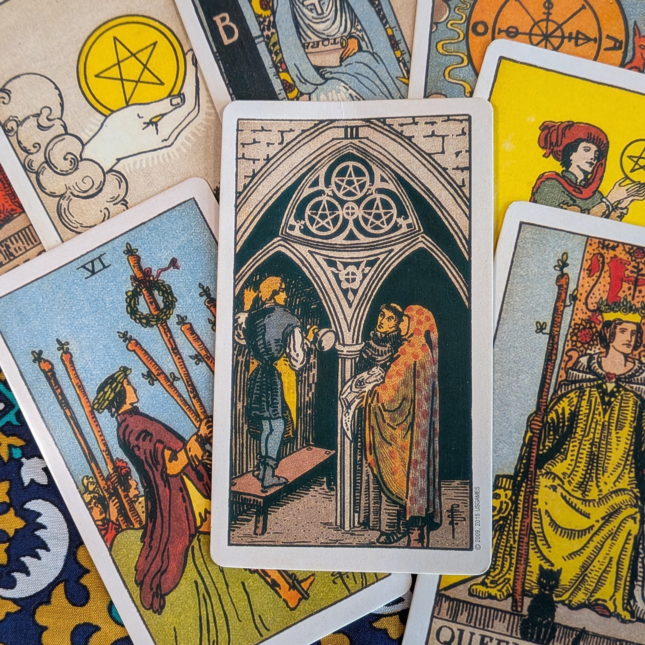 A close-up photograph of Rider-Waite-Smith style tarot cards spread across a patterned surface. The Three of Pentacles card is centred, showing three figures beneath a gothic arch, surrounded by other visible cards including the Six of Wands, Ace of Pentacles, Queen of Wands and Page of Pentacles. The image is used to promote free community partner audio spots for Tarot Interviews, open to tarot, folklore, heritage, arts and culture organisations.
