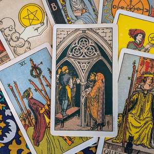 A close-up photograph of Rider-Waite-Smith style tarot cards spread across a patterned surface. The Three of Pentacles card is centred, showing three figures beneath a gothic arch, surrounded by other visible cards including the Six of Wands, Ace of Pentacles, Queen of Wands and Page of Pentacles. The image is used to promote free community partner audio spots for Tarot Interviews, open to tarot, folklore, heritage, arts and culture organisations.