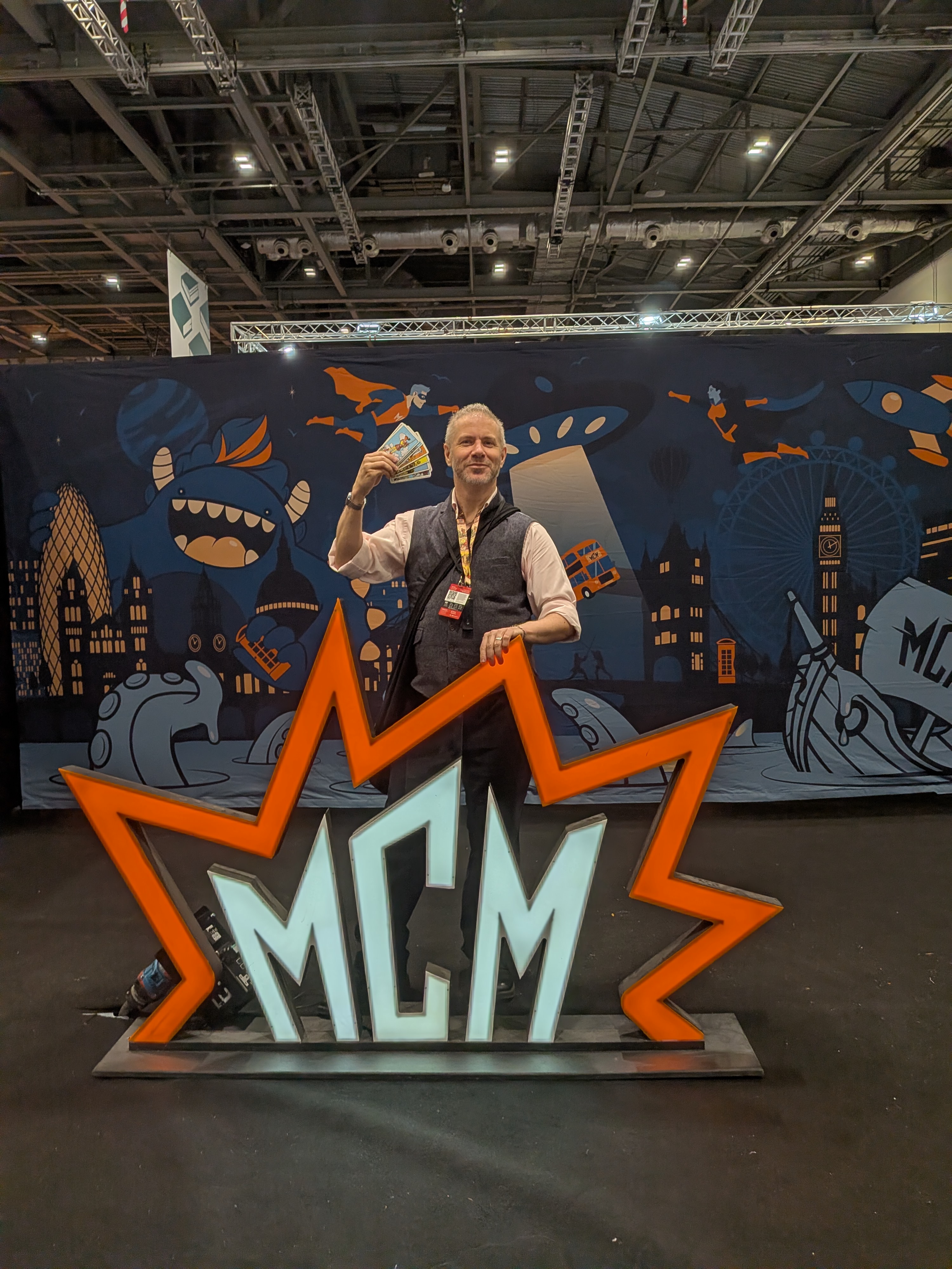 Finbarre Snarey poses behind a large orange and white MCM Comic Con sign at ExCeL London, holding up a fan of Rider–Waite–Smith tarot cards. Wearing a waistcoat and press lanyard, he stands against a colourful illustrated backdrop featuring fantastical creatures, superheroes and London landmarks during MCM London Comic Con, October 2026.