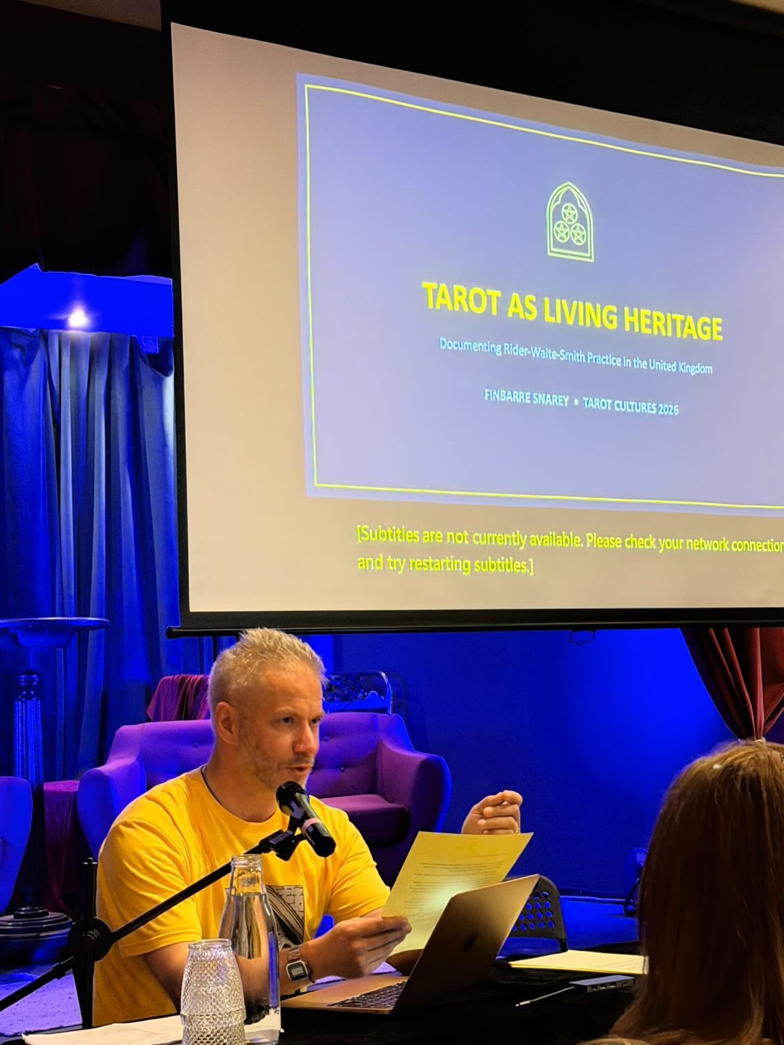 Finbarre Snarey sits at a conference table with a microphone, laptop and printed notes while presenting “Tarot as Living Heritage” at Tarot Cultures 2026. Behind him, a large projected slide shows the title of the talk and the subtitle “Documenting Rider-Waite-Smith Practice in the United Kingdom.” The room is lit with blue stage lighting.