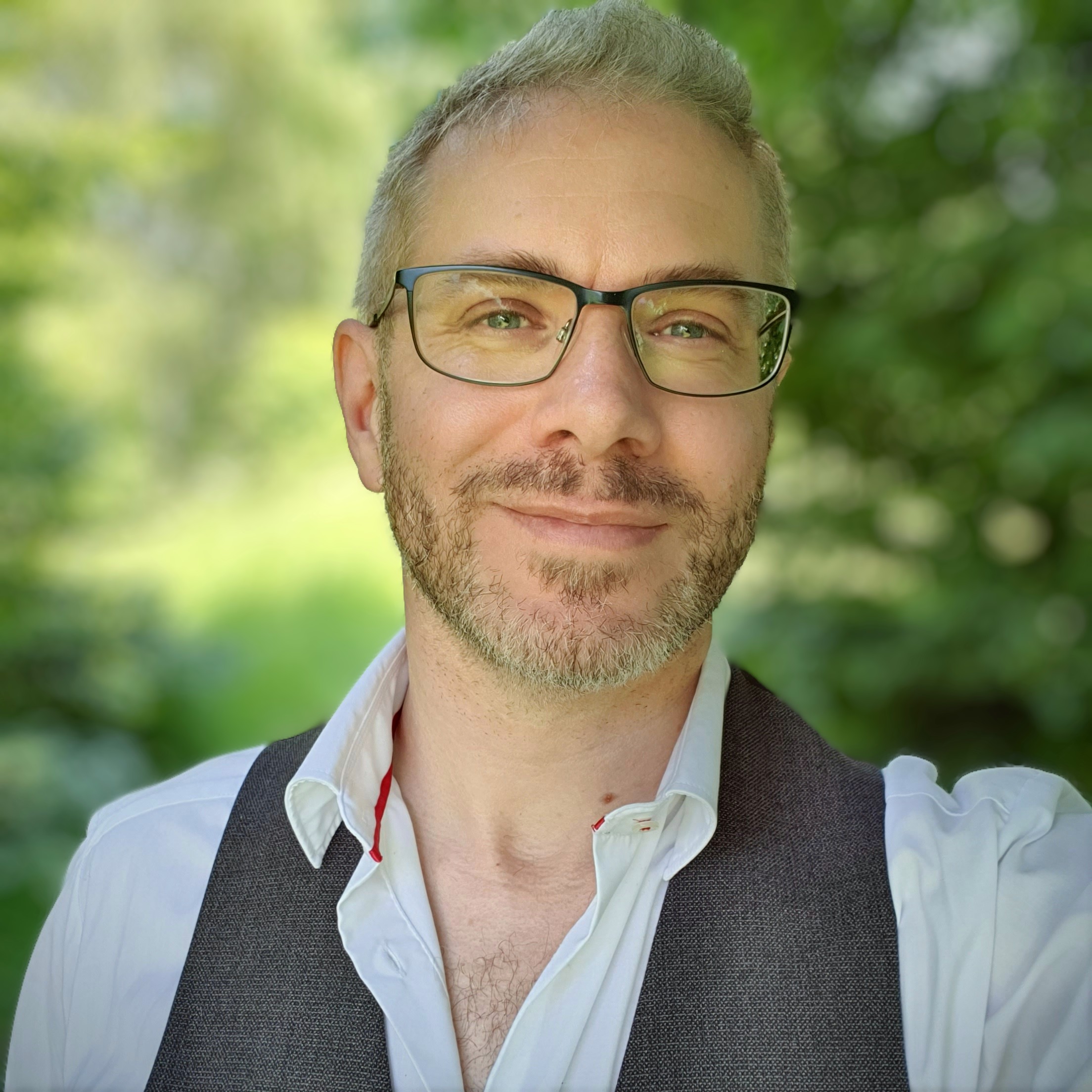 Portrait of Finbarre Snarey smiling outdoors, wearing glasses, a white shirt and grey waistcoat, against a softly blurred green background.